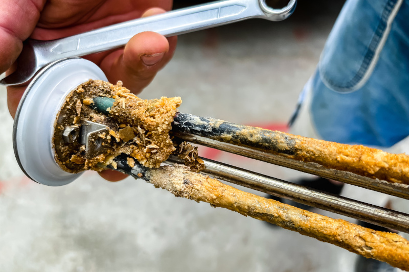 Water Tank heating element that is heavily coated with mineral deposits and scale buildup, likely due to hard water. The corrosion and deposits reduce the efficiency of the heating element and can eventually lead to failure if not cleaned or replaced.